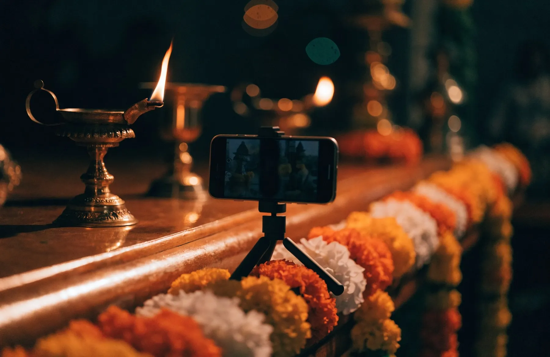 A phone on a small tripod filming a brass aarti lamp burning beside marigold garlands on the altar ledge