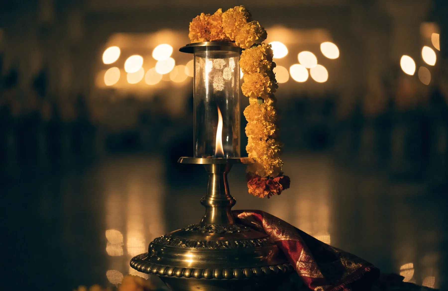 An akhand jyot burning inside a tall glass chimney on a brass stand, dressed with a marigold garland, with lamps glowing behind