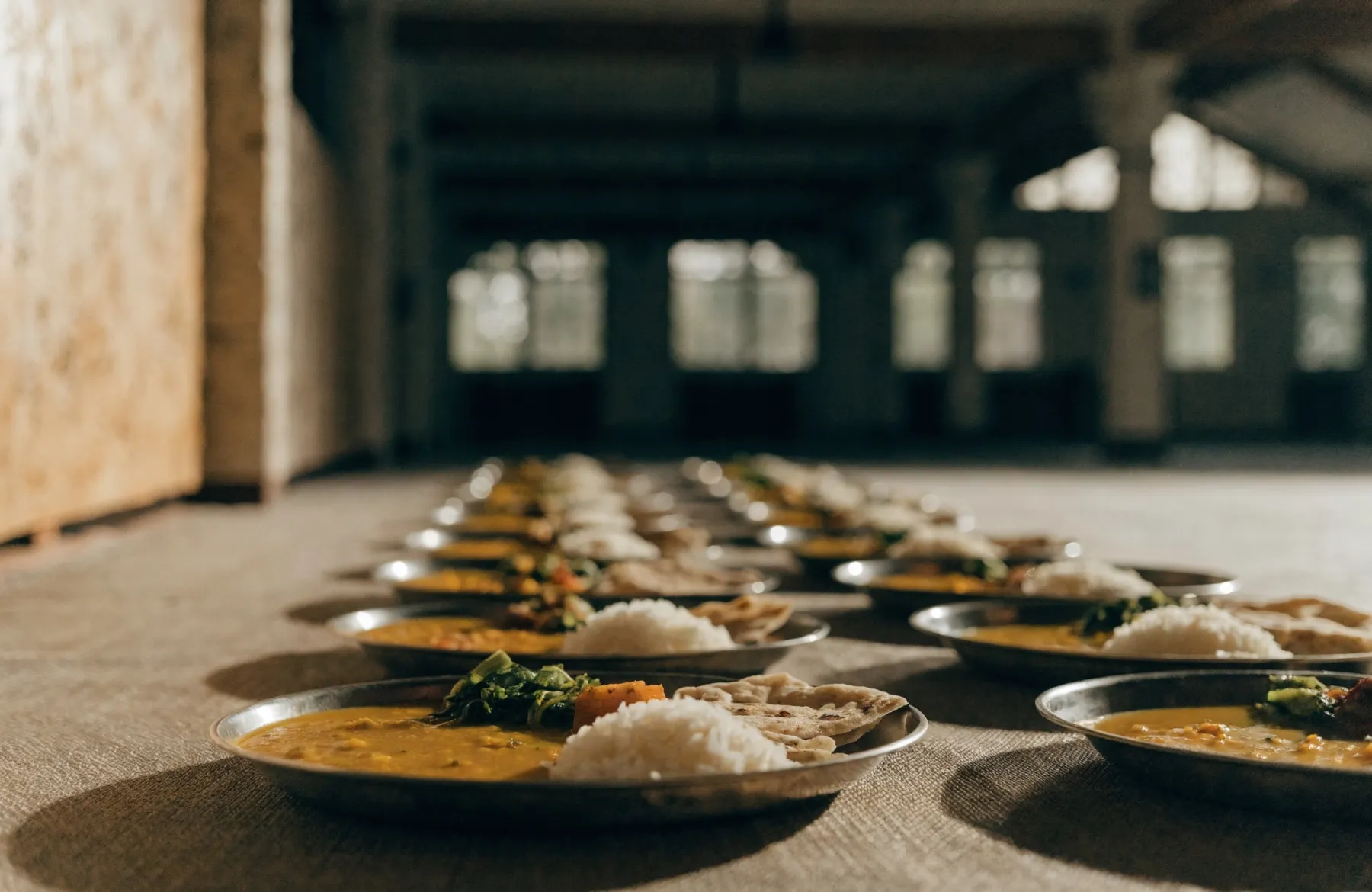 Rows of steel thali plates filled with dal, rice, roti and vegetables, laid out along the floor of a hall