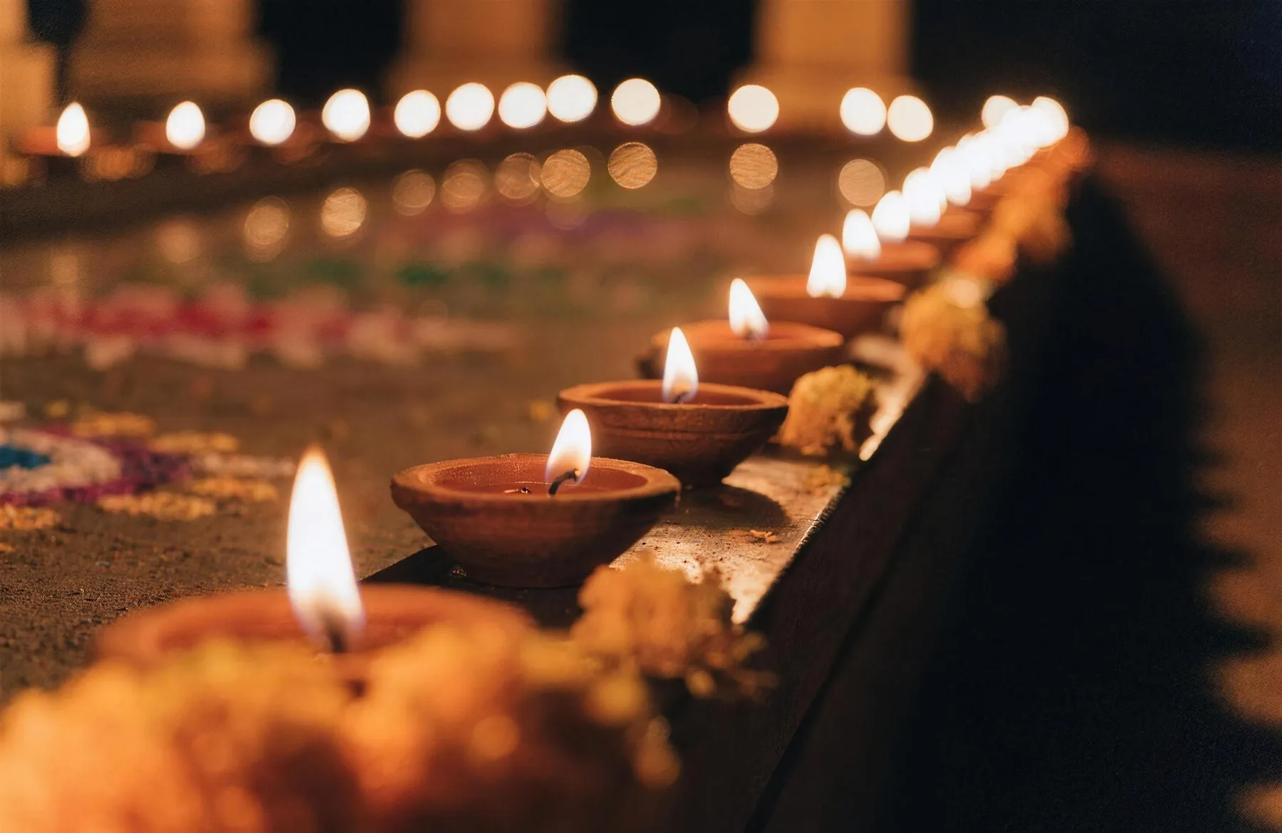 A row of small clay diya lamps burning along a ledge beside marigolds and rangoli