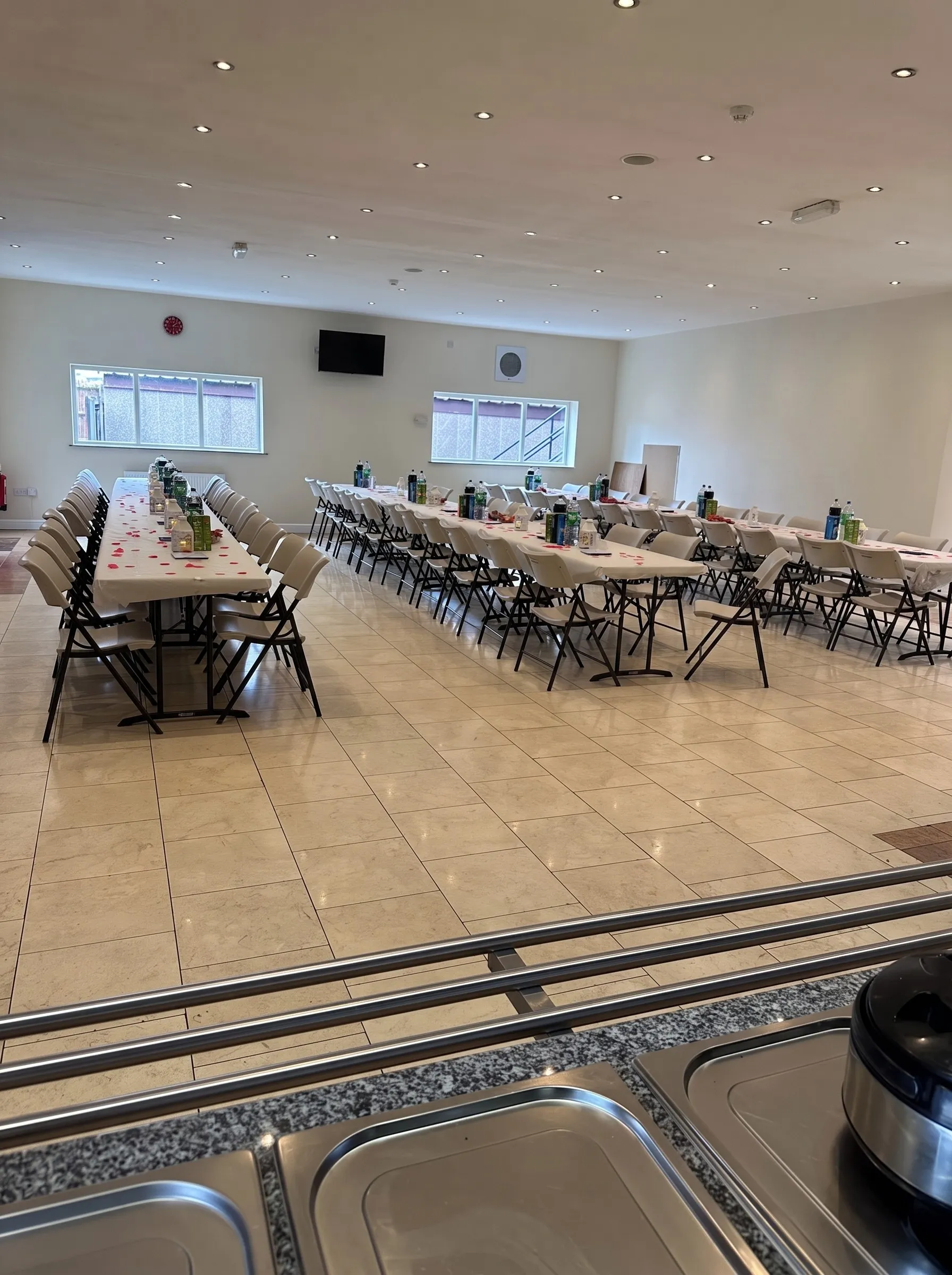 The mandir's function hall set for an event — long tables in white cloths with folding chairs, rose petals scattered along them, and the serving counter in the foreground