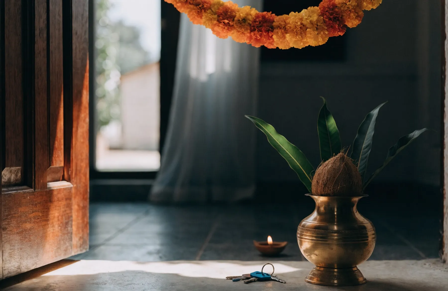 A kalash and a lit diya on the threshold of a new home, house keys on the floor and a marigold toran hanging above the open door