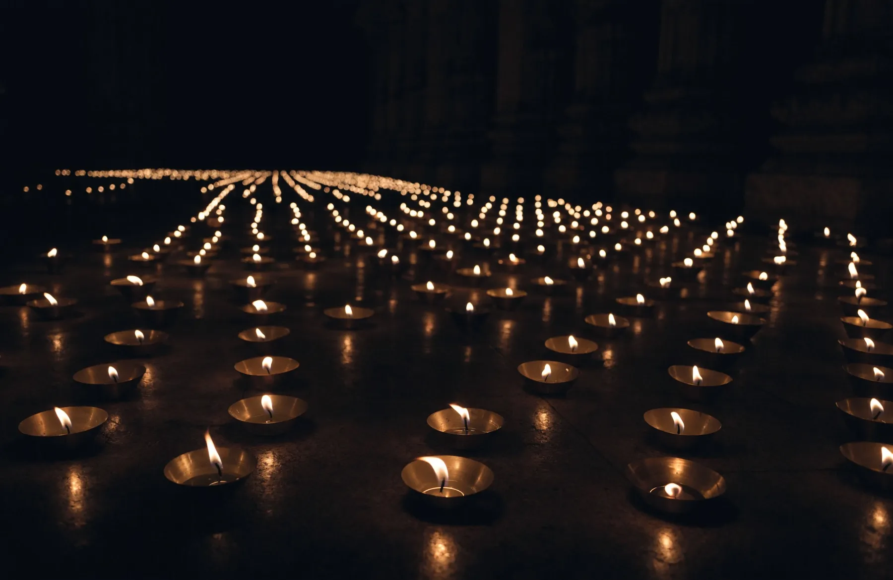 Many small brass oil lamps burning across a temple floor, receding into the darkness of the new-moon night