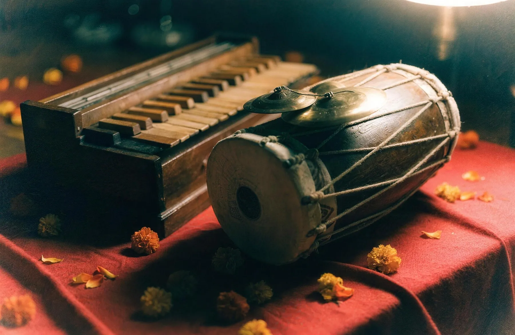 A dholak, a harmonium and brass manjira cymbals resting on a red cloth in warm lamplight