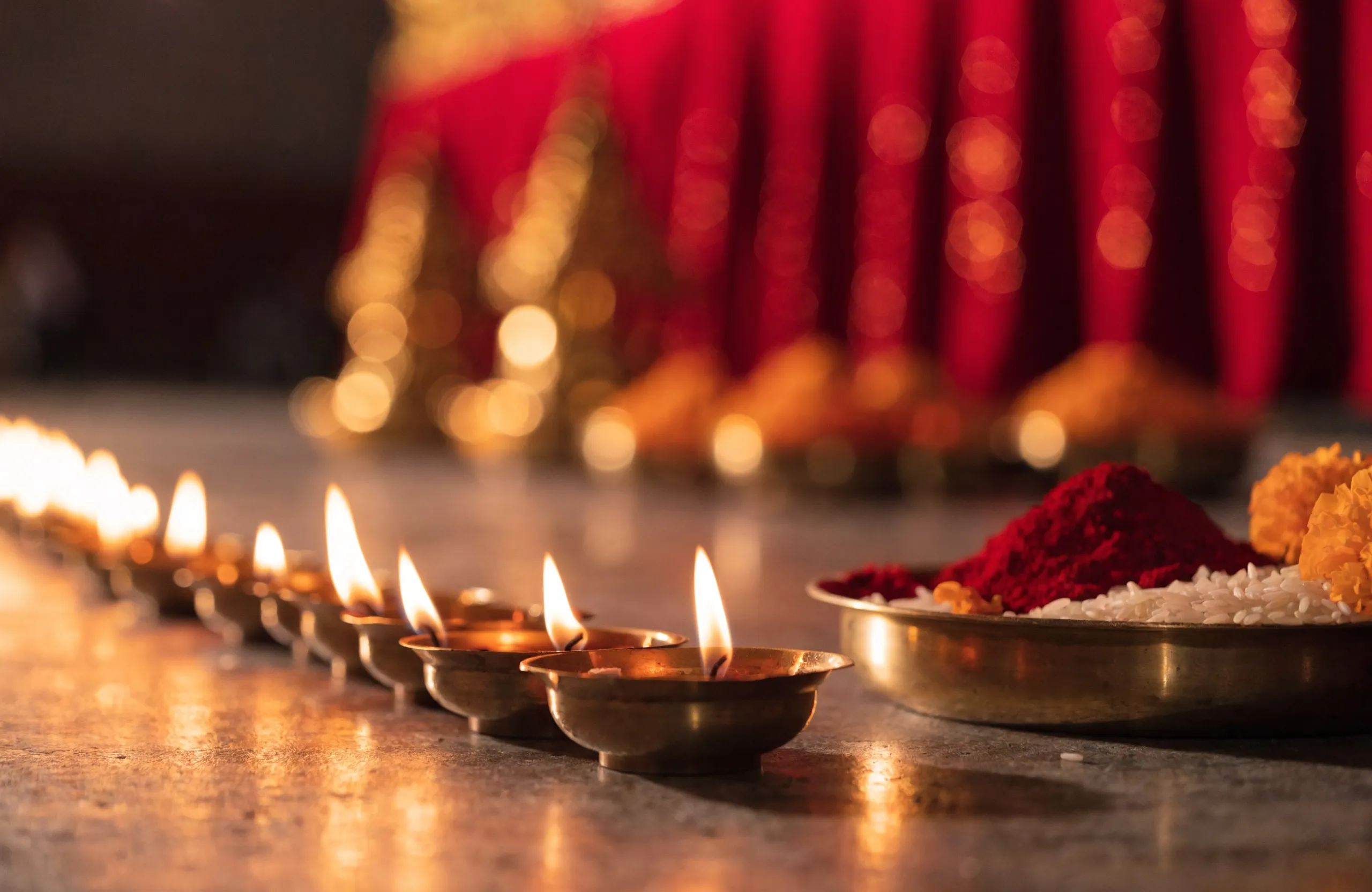 A row of lit brass diyas beside a thali of kumkum, rice and marigolds