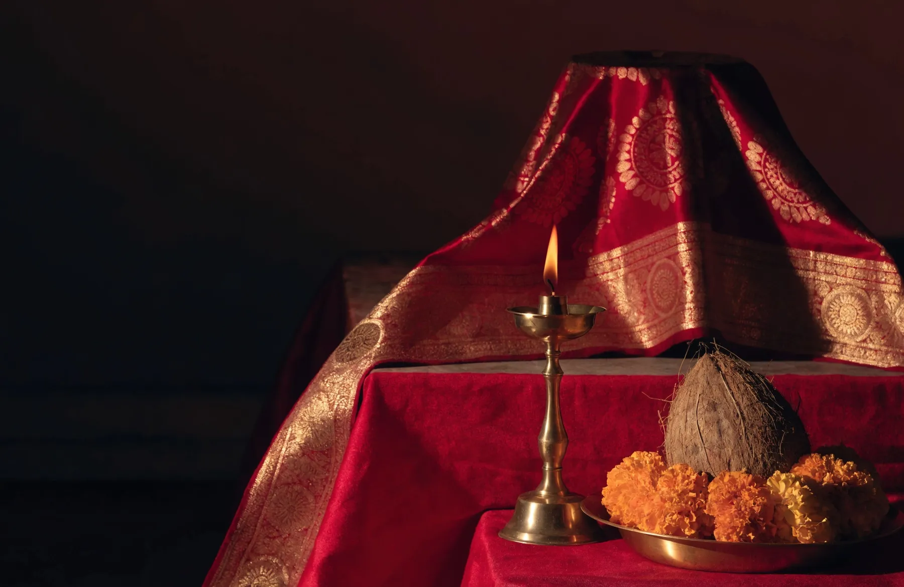 Maa's jyot burning before a crimson and gold chunni, with marigolds and a coconut on a brass thali