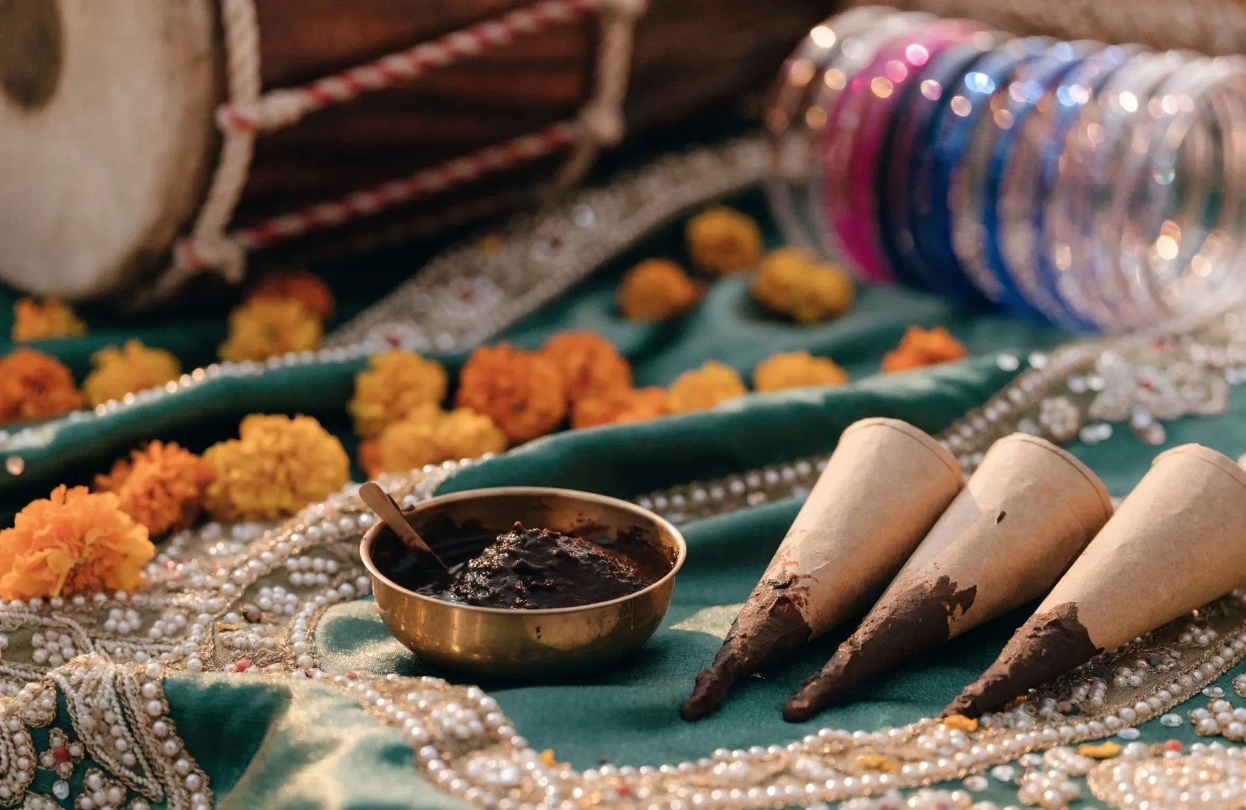 Mehndi cones and a bowl of henna paste on embroidered green and gold fabric, with bangles and a dholak behind