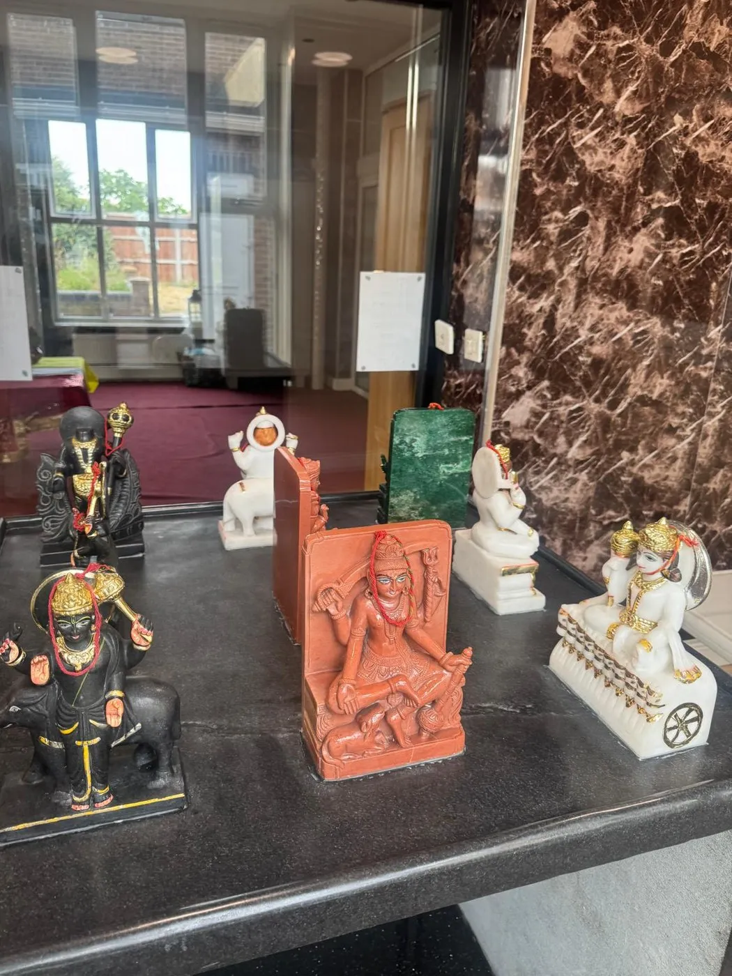 The Navagraha shrine at the mandir — the nine grahas as separate small murtis on a black plinth, Shani in black upon his buffalo, Mangal in terracotta and Surya on his chariot wheel in white marble