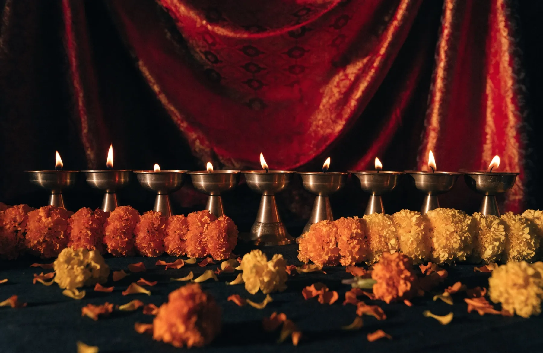 Nine brass oil lamps burning in a row before red and gold cloth, with marigold garlands before them