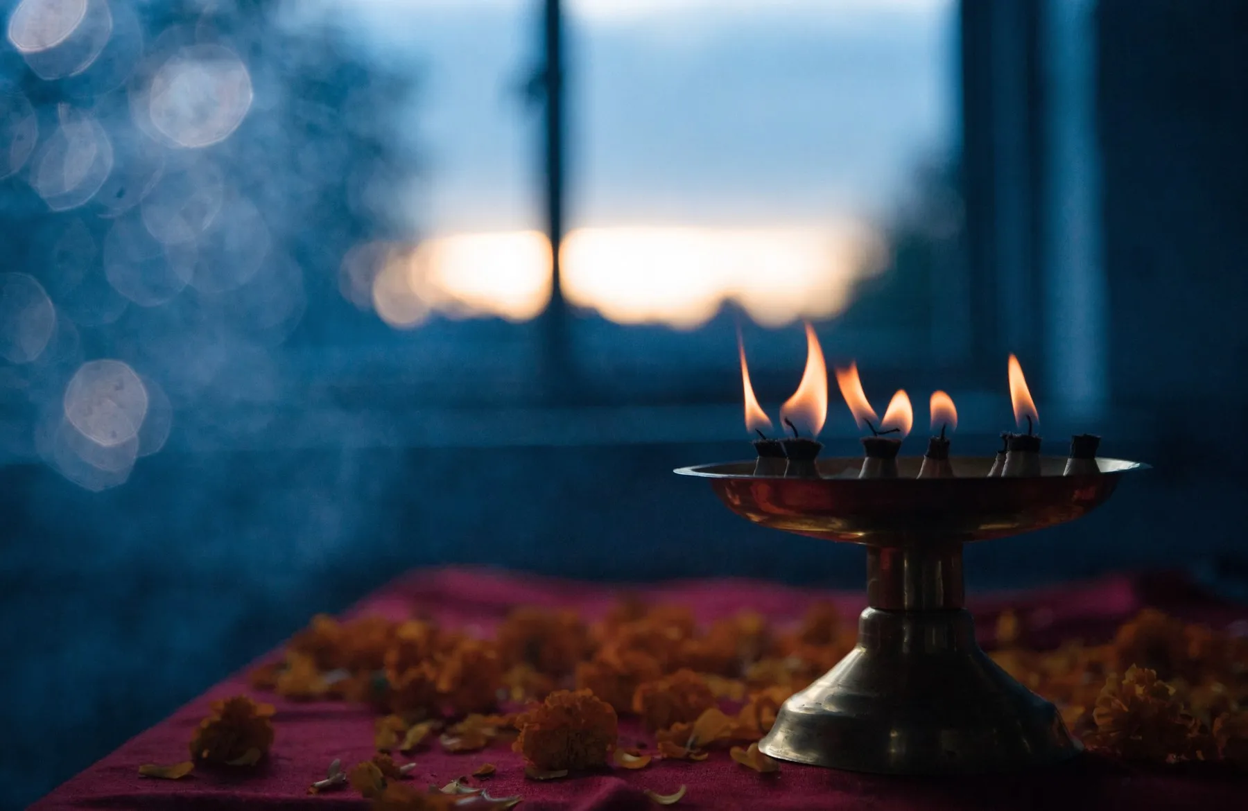 A brass aarti lamp with several wicks burning as pale gold dawn light breaks through a window behind it