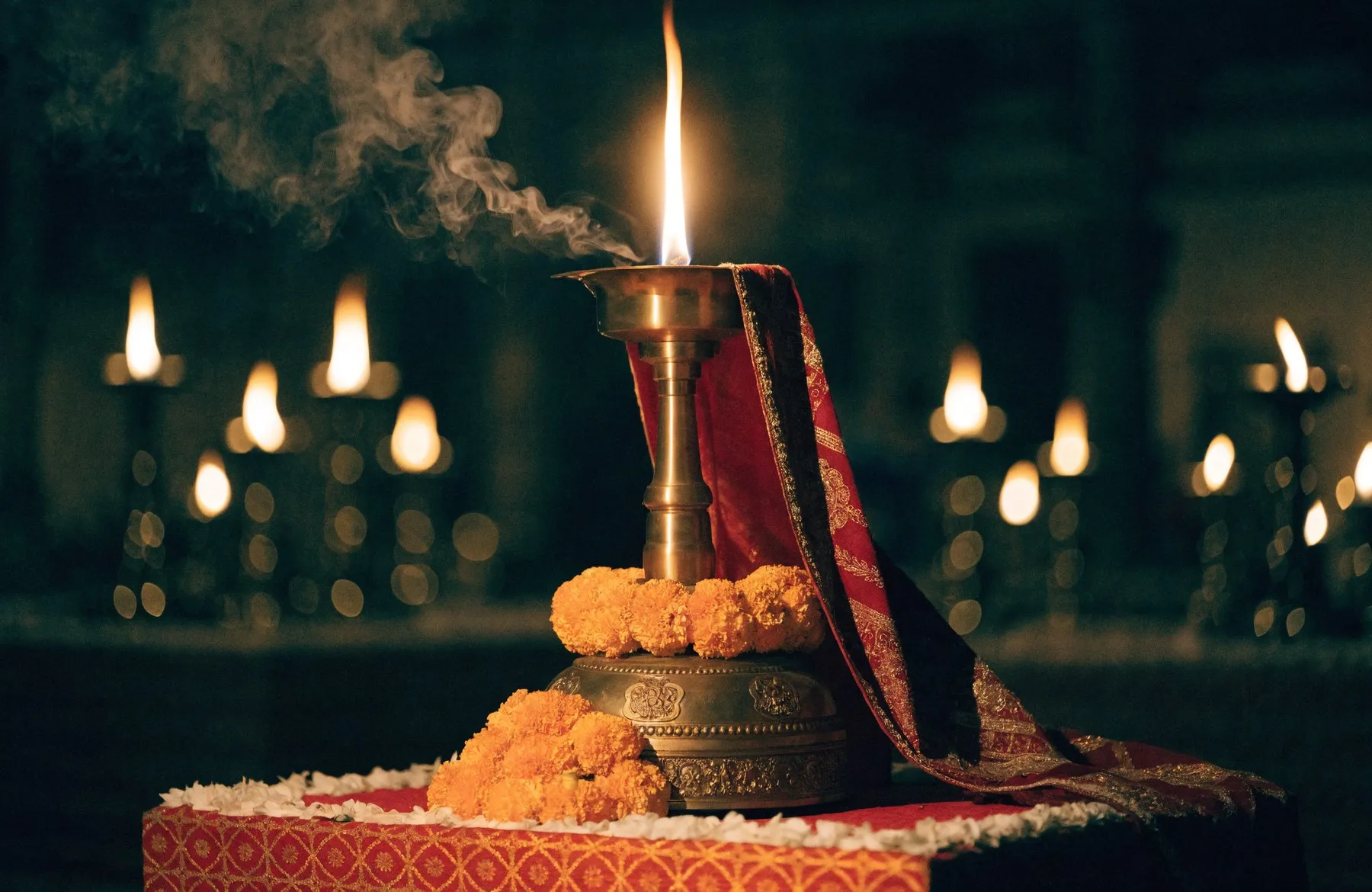 A tall golden flame burning upward from an ornate brass jyoti lamp dressed in red and gold cloth, marigolds at its base