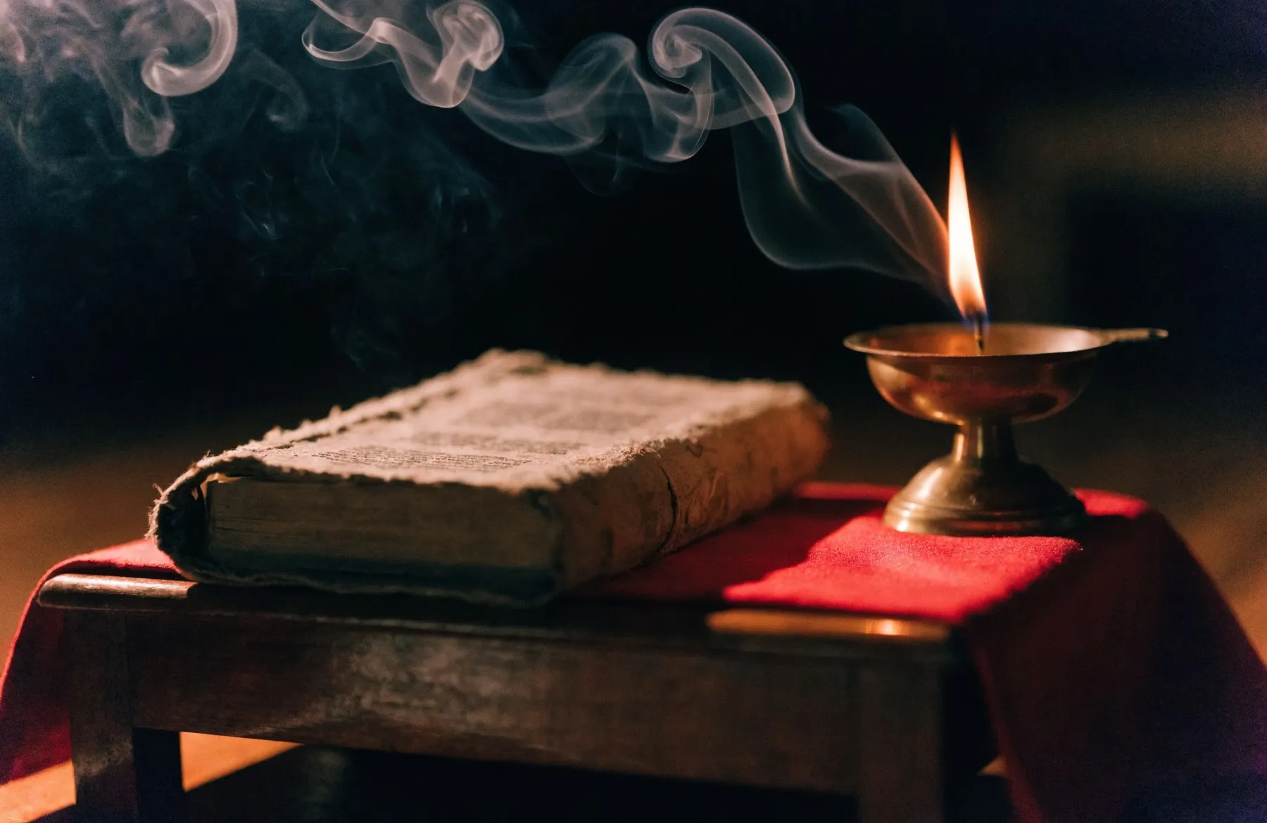 An open scripture resting on a red cloth-covered stand with a small oil lamp burning beside it in the dark