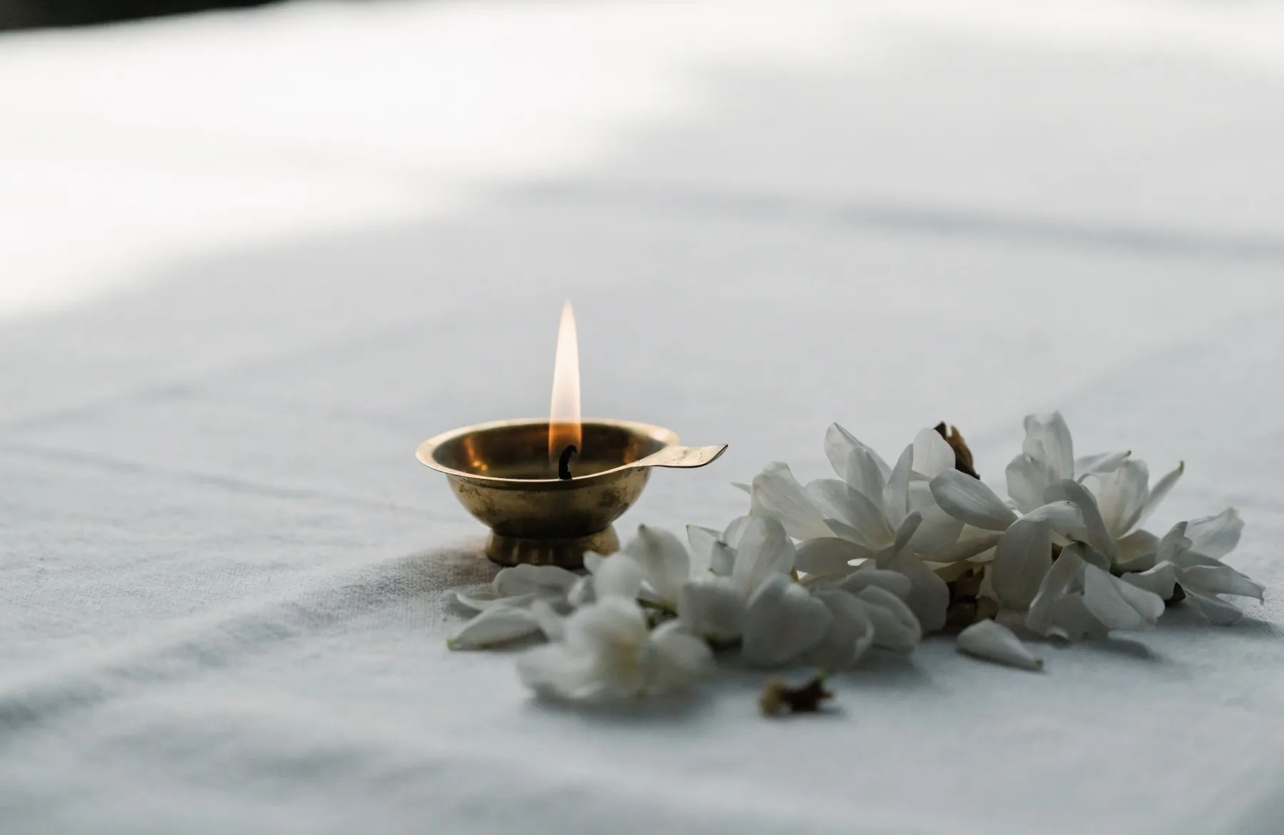 A single brass lamp burning quietly beside white jasmine flowers on plain white cloth