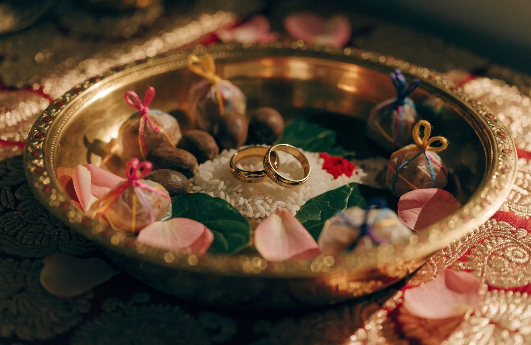 A decorated brass thali holding two gold rings, rice, kumkum, betel leaves and wrapped sweets on red and gold silk — a sagai ceremony