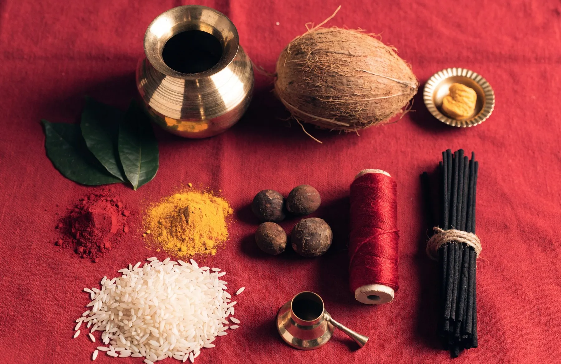 Pooja samagri laid out on a red cloth — a brass kalash, coconut, betel leaves and nuts, rice, kumkum and turmeric, red mauli thread, incense and a diya