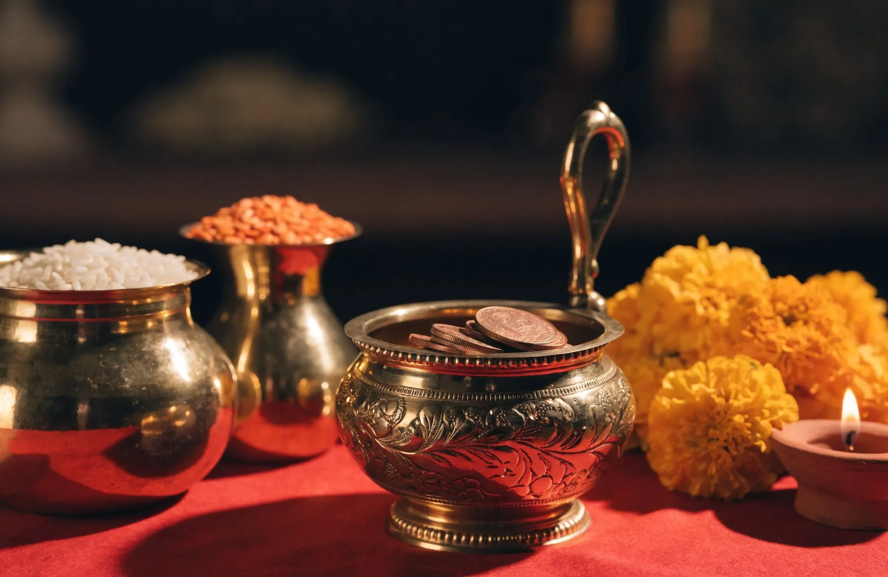 Brass vessels of rice and lentils beside a brass daan bowl with coins, marigolds and a lit diya