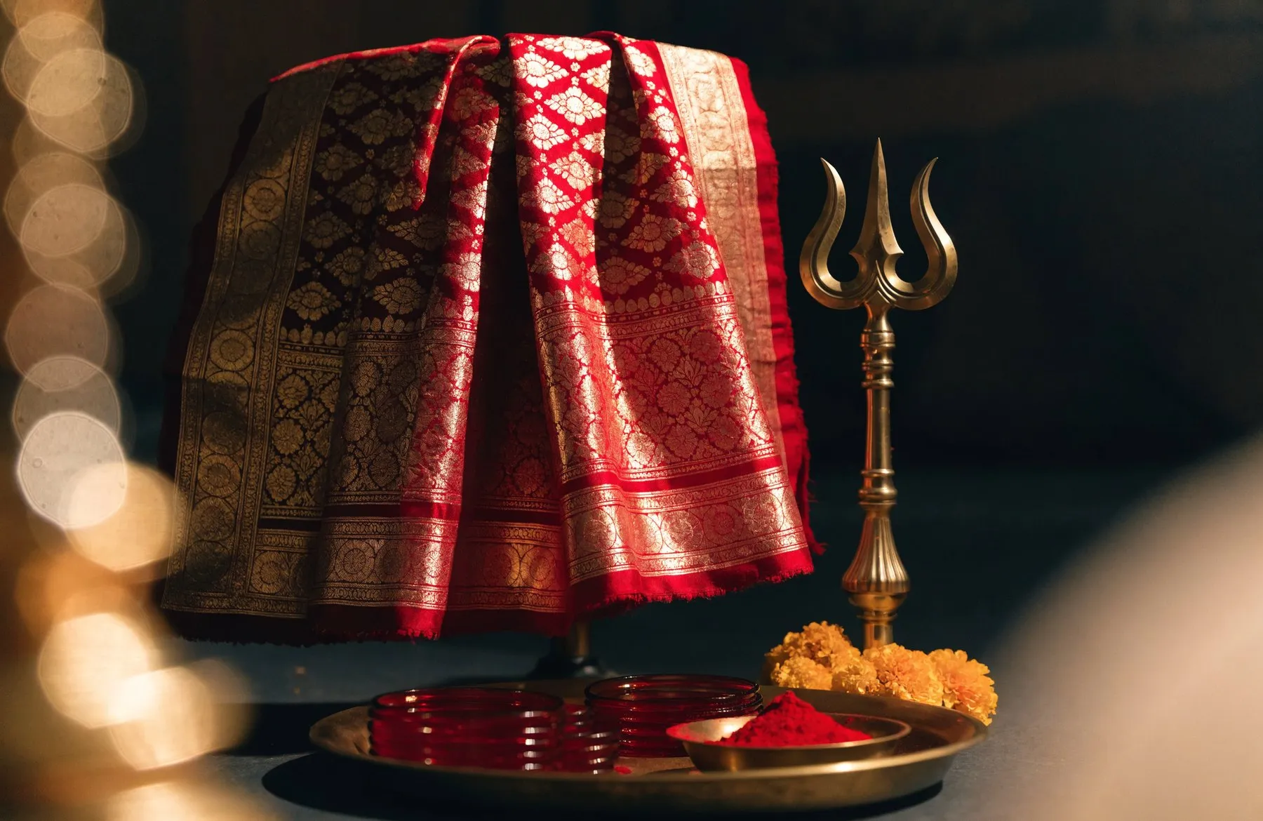 A red and gold chunri draped beside a brass trishul, with glass bangles and sindoor on a thali below