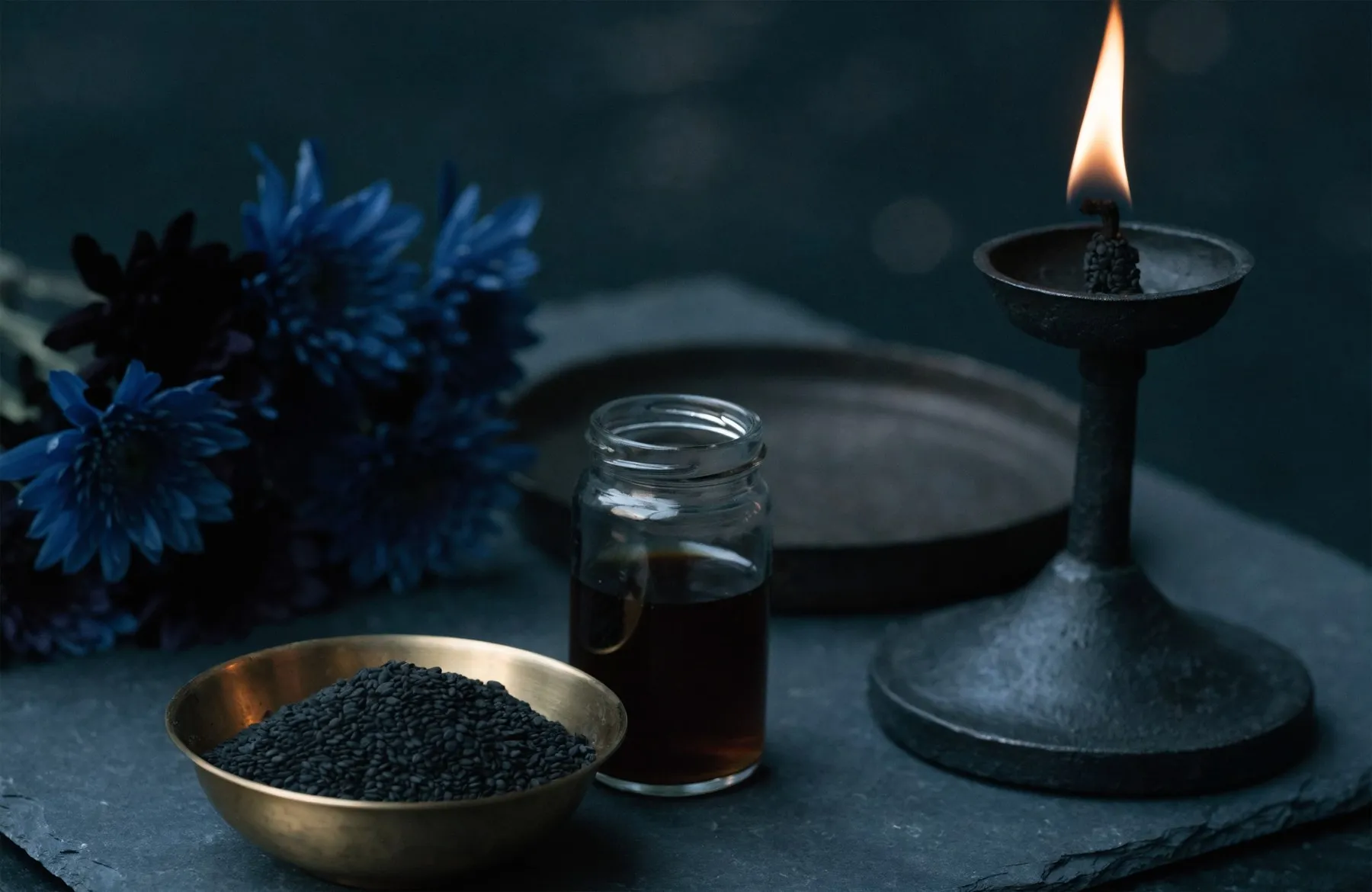 Offerings for Shani Dev on dark slate — a brass bowl of black sesame seeds, a jar of mustard oil, an iron dish, deep blue flowers and an iron lamp burning