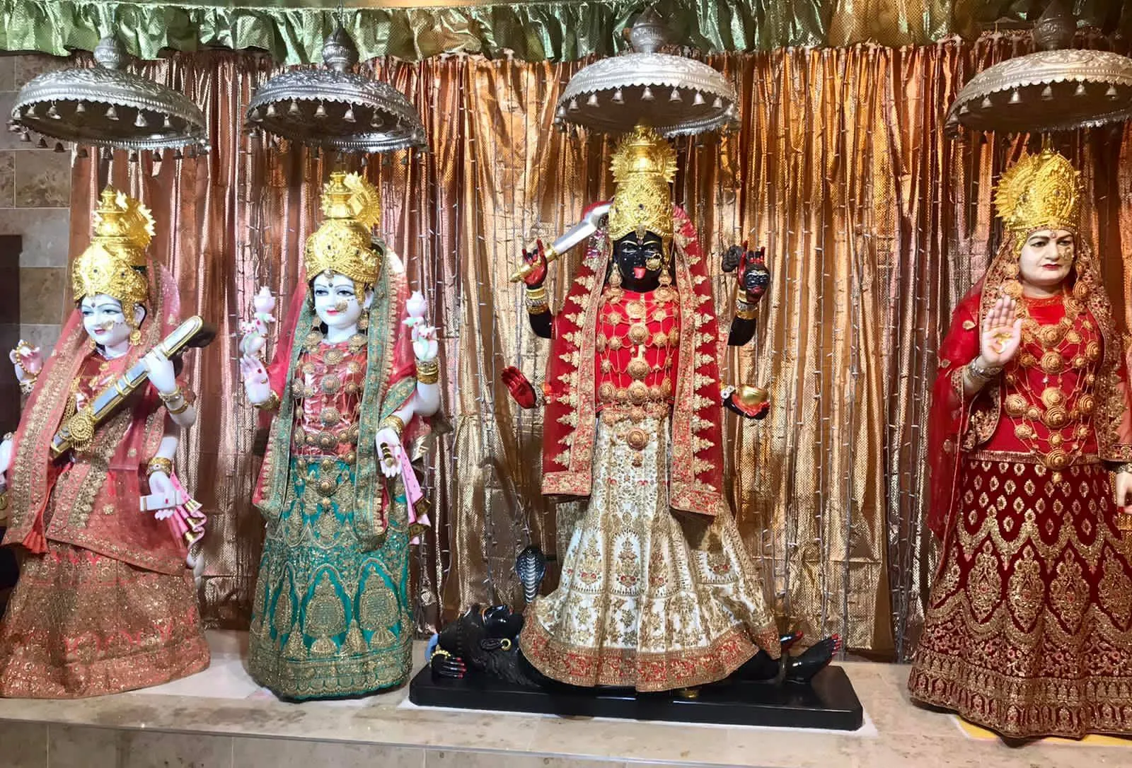 The altar before a copper-coloured drape, Maa Saraswati in pink, Maa Lakshmi in teal green, Maa Kali in red and Maa Bhameshwari in deep red