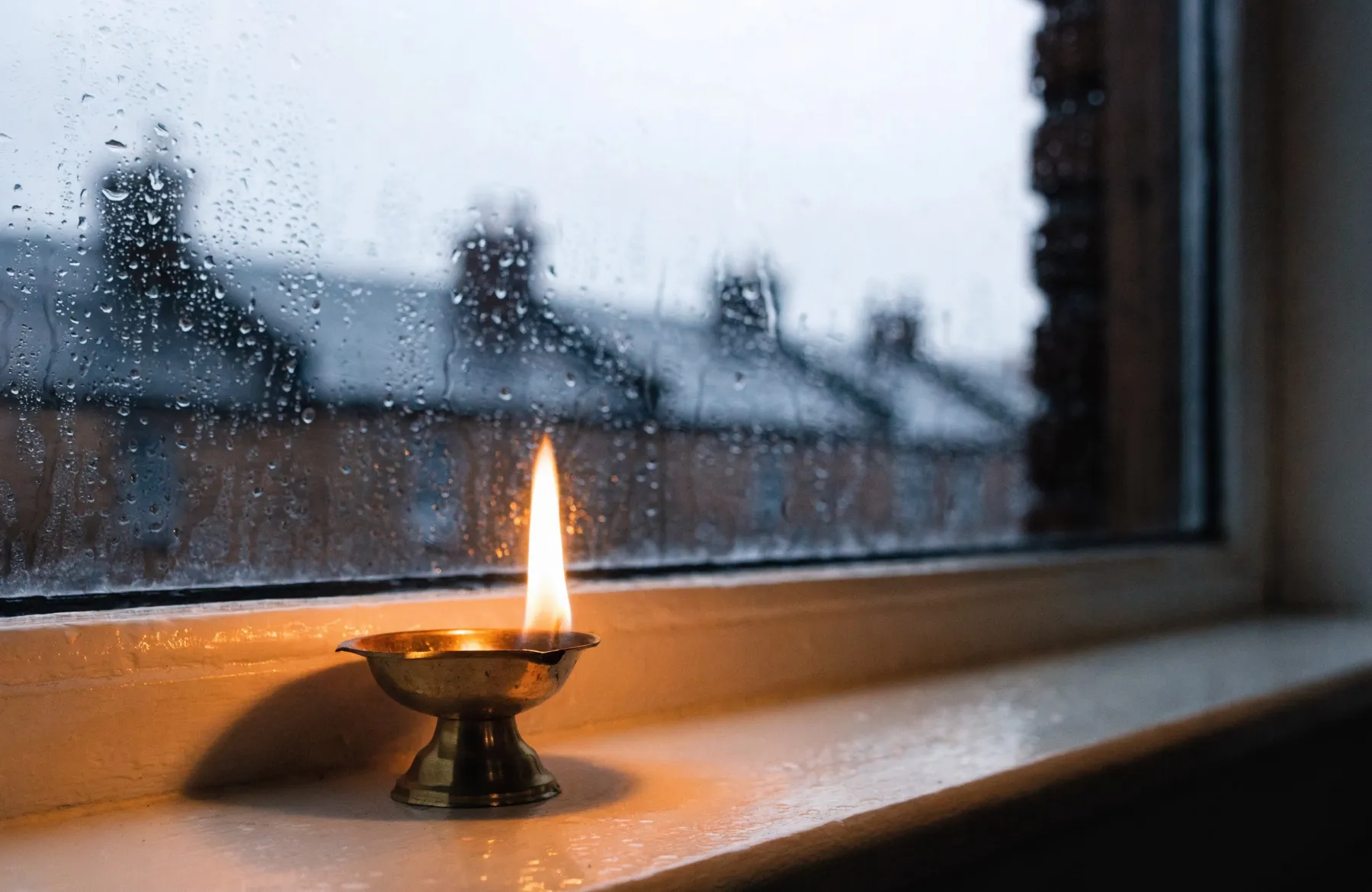 A brass diya burning on a windowsill against a rain-streaked window and a grey British sky