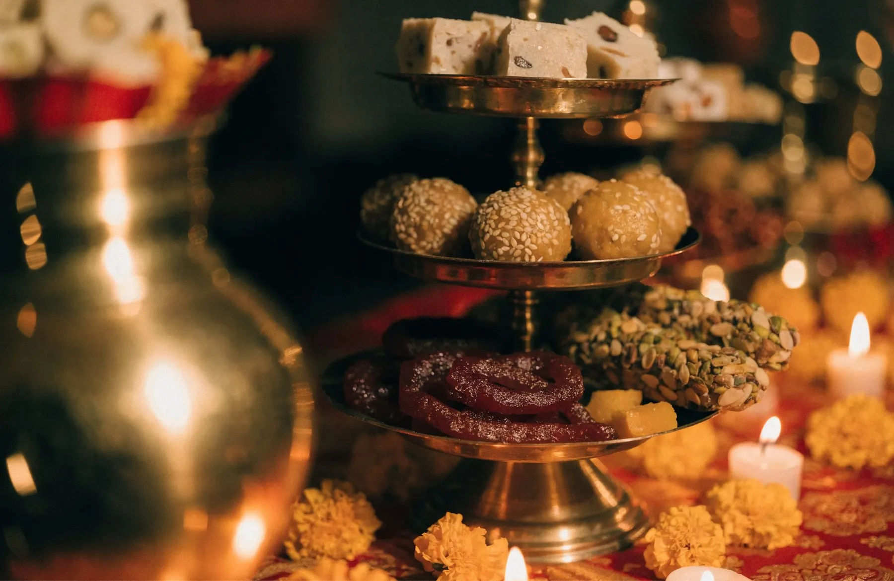 A tiered brass stand of Indian mithai — barfi, ladoo and jalebi — with candles and marigold petals around it