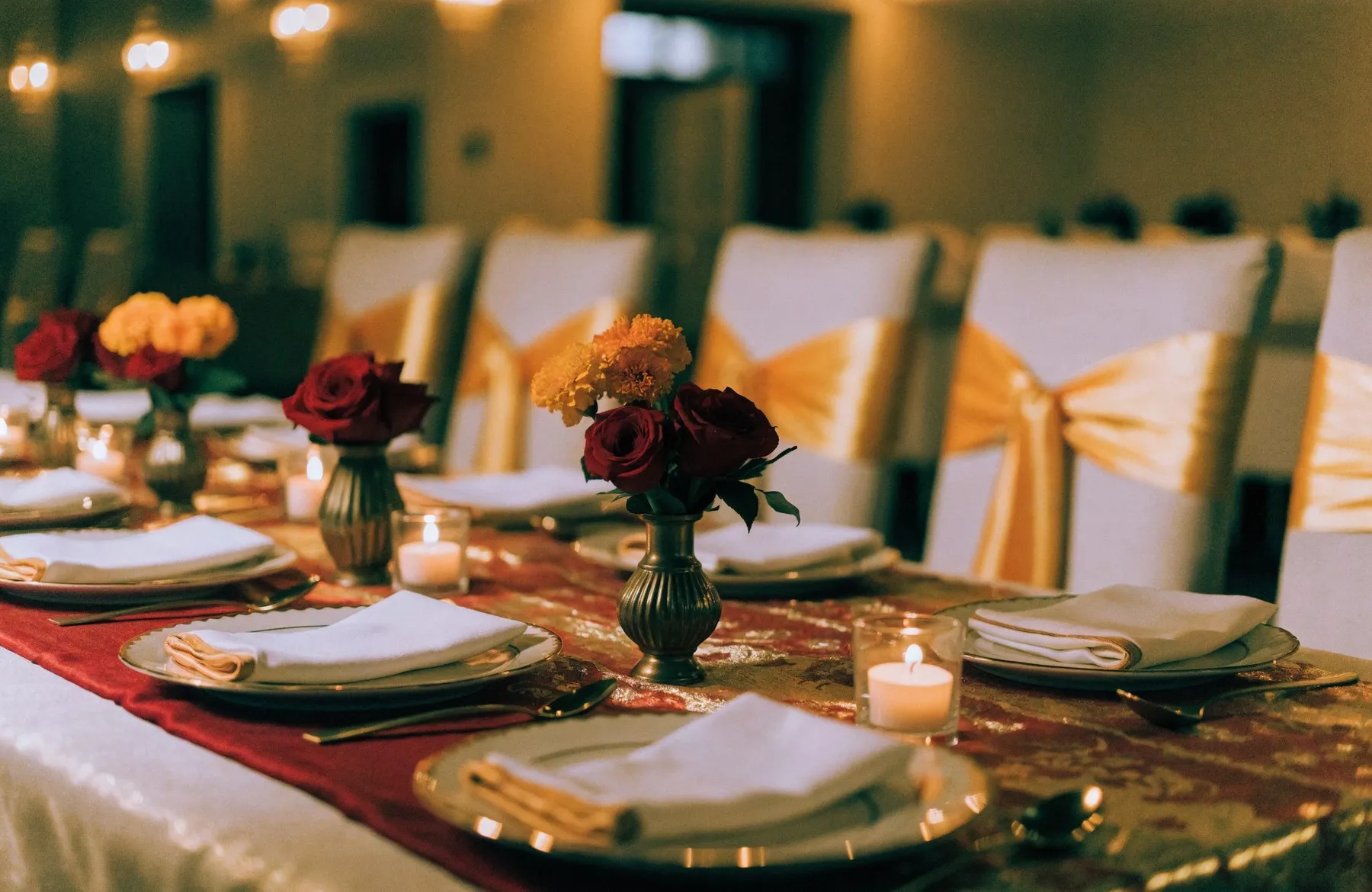 A wedding reception table dressed in red and gold with roses, brass vases and candles, chairs with gold sashes behind