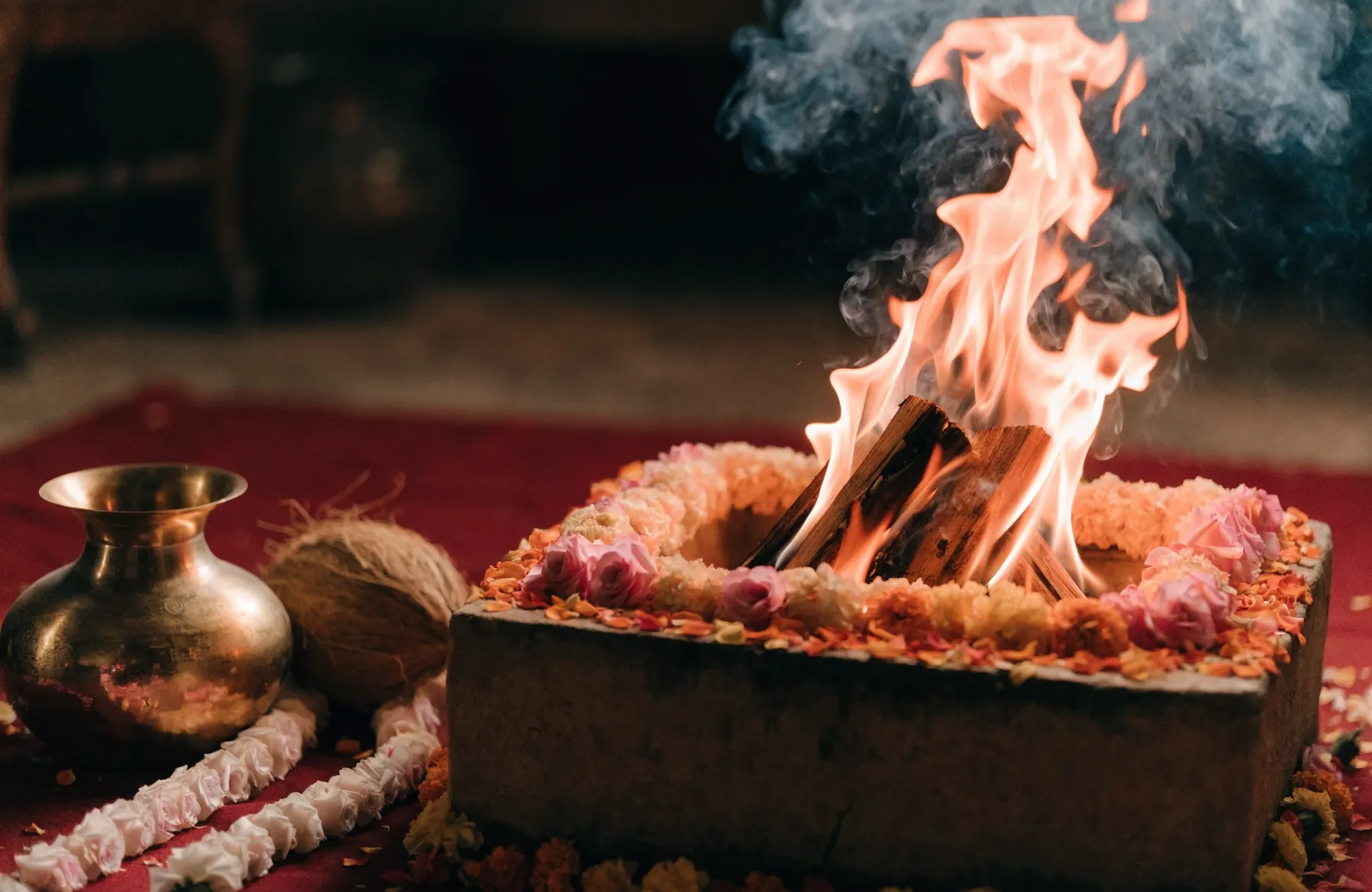 A Hindu wedding sacred fire burning in a havan kund, ringed with marigold and rose petals, with a brass kalash and garlands beside it