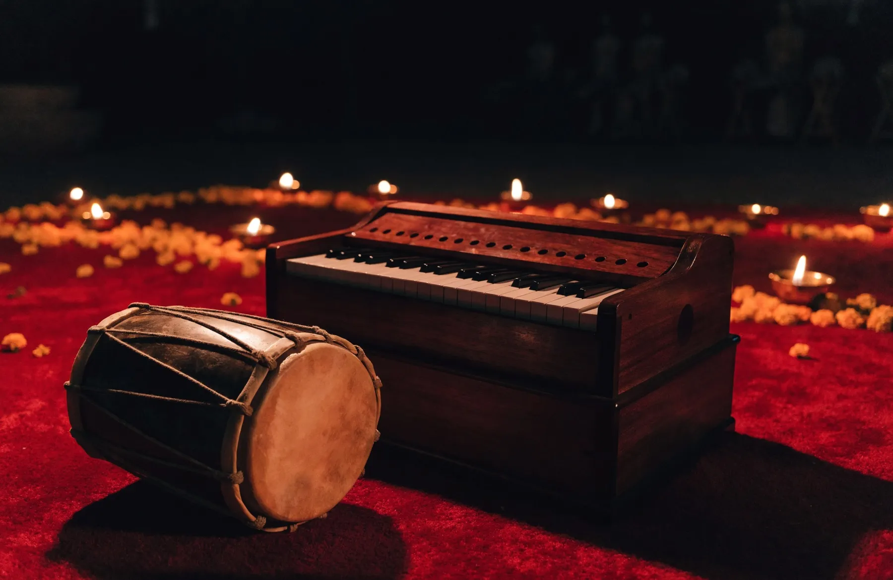 A harmonium and dholak resting on a red carpet at night, lit by oil lamps, before an all-night gathering begins
