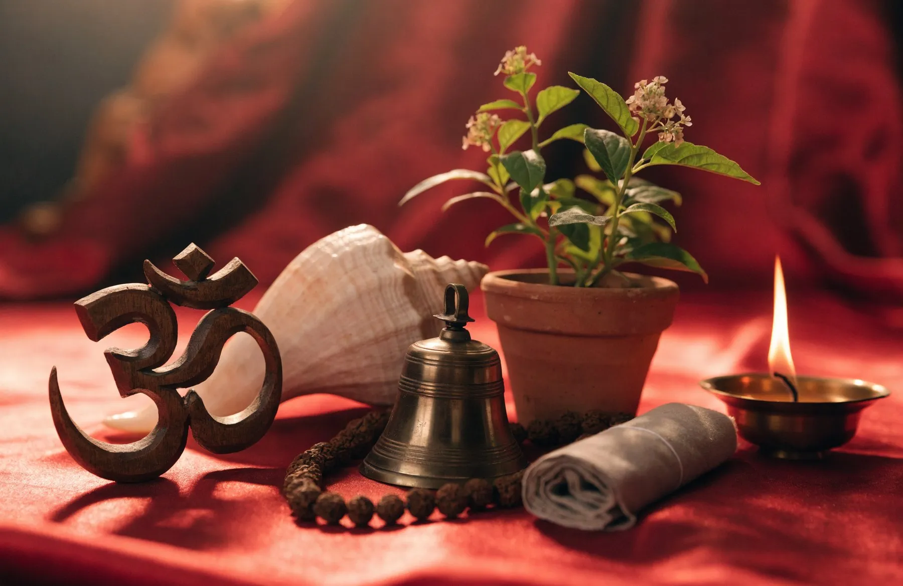 Devotional objects together on red silk — a carved Om, a conch, a bell, a tulsi plant, cloth-wrapped scripture, a lamp and a rudraksha mala