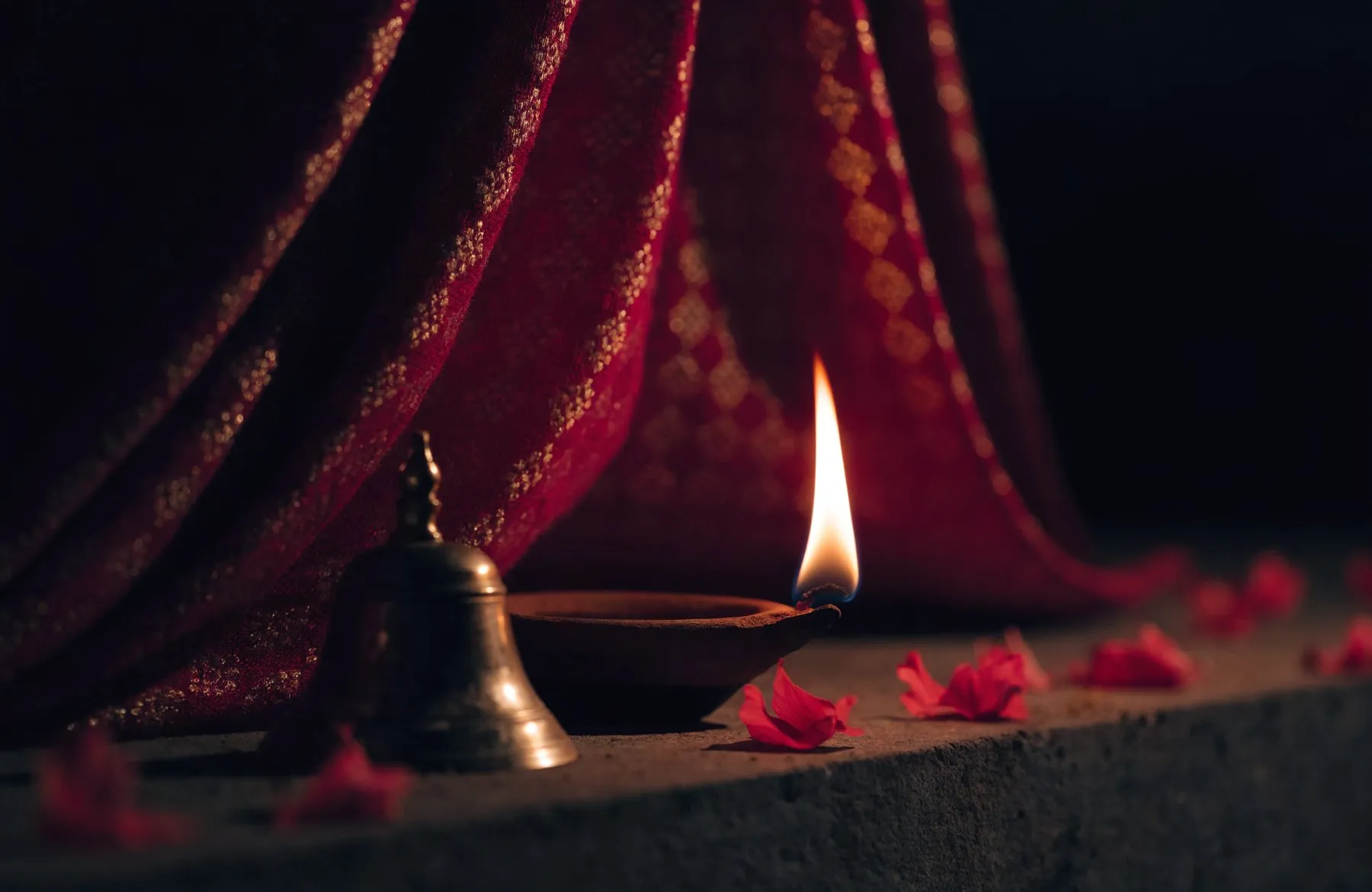 A single oil lamp burning before a deep crimson and gold chunri in near darkness, with a brass bell and red hibiscus petals on the ledge below