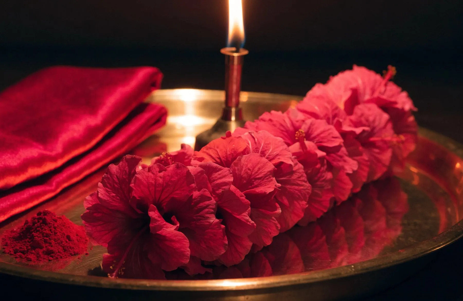 A long garland of deep red hibiscus coiled on a brass thali, a tall oil lamp burning behind it
