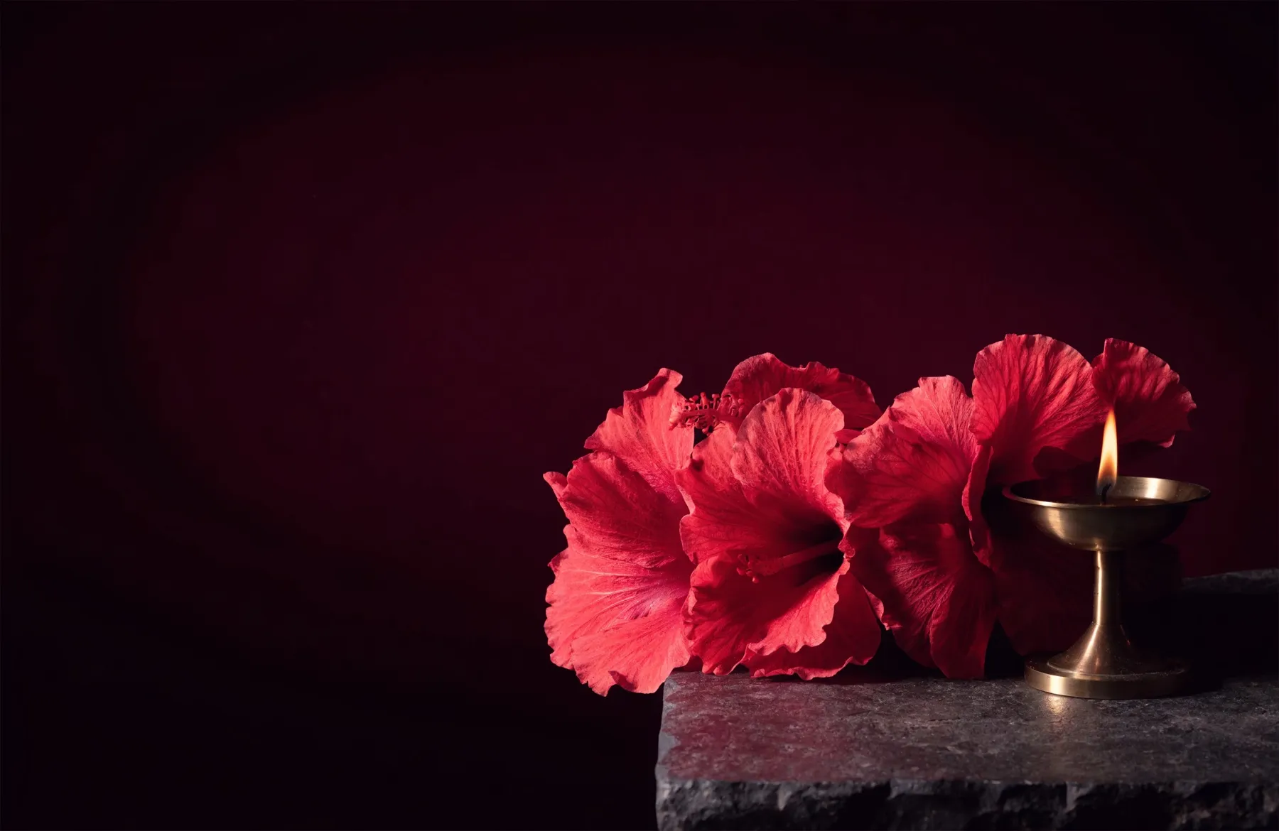 A garland of deep red hibiscus flowers — the flower most loved by Maa Kali — beside a burning brass diya in the dark