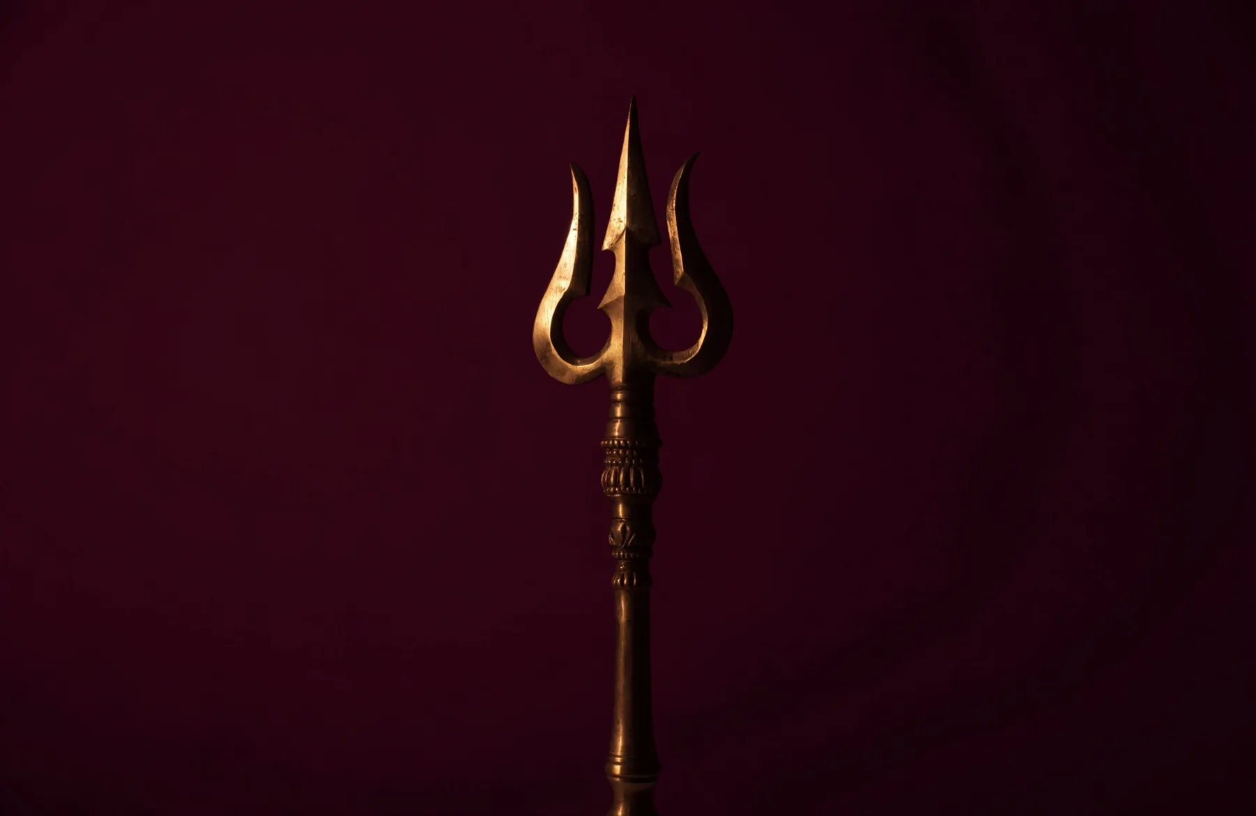 A brass trishul standing upright in a single shaft of golden light against deep maroon darkness